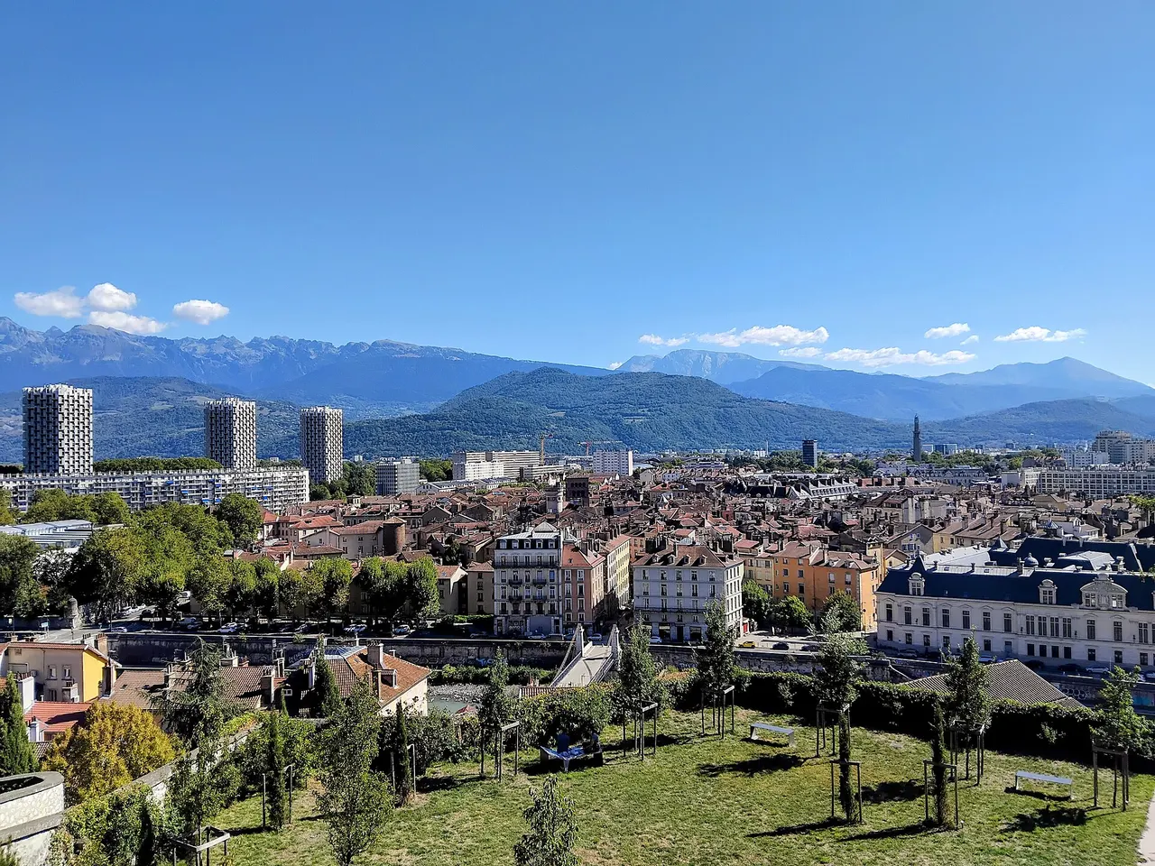 Les toits de Grenoble et les trois tours de l’Île Verte vus depuis les hauteurs, avec les montagnes alpines à l’horizon sous un ciel bleu.