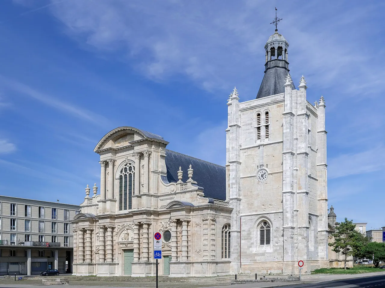 La cathédrale Notre-Dame du Havre, sa façade à colonnes et sa haute tour-clocher blanche sous un ciel bleu, un immeuble de la reconstruction à gauche.