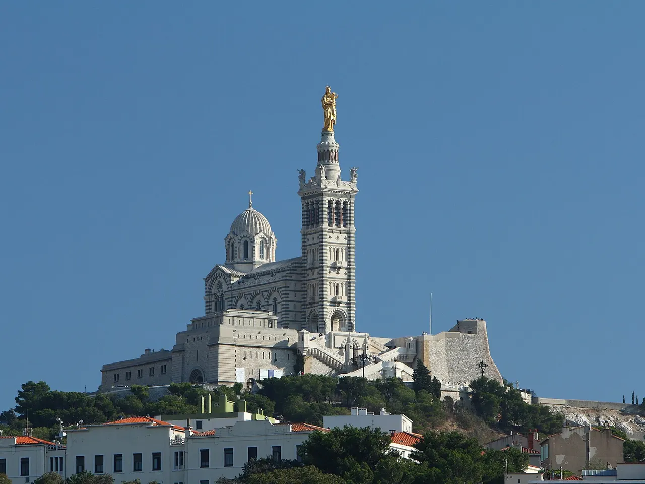 La basilique Notre-Dame-de-la-Garde et sa Vierge dorée sur leur colline, au-dessus des toits de Marseille, sous un ciel bleu.