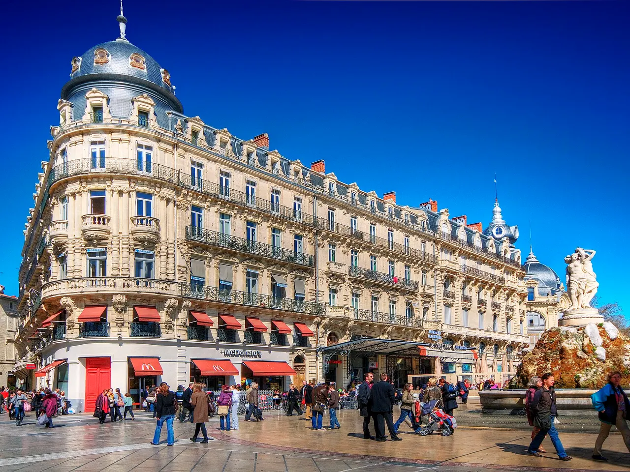 Immeuble à dôme, fontaine des Trois Grâces et passants sur la place de la Comédie, à Montpellier, sous un ciel bleu.