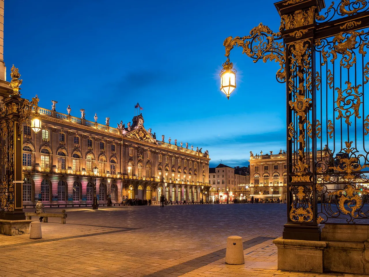 Place Stanislas à Nancy au crépuscule, l’hôtel de ville illuminé vu à travers les grilles dorées de Jean Lamour
