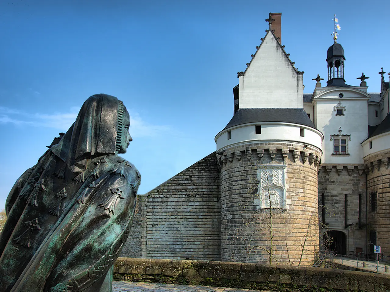 Statue de bronze d’Anne de Bretagne devant une tour du château des ducs de Bretagne, à Nantes, sous un ciel bleu.