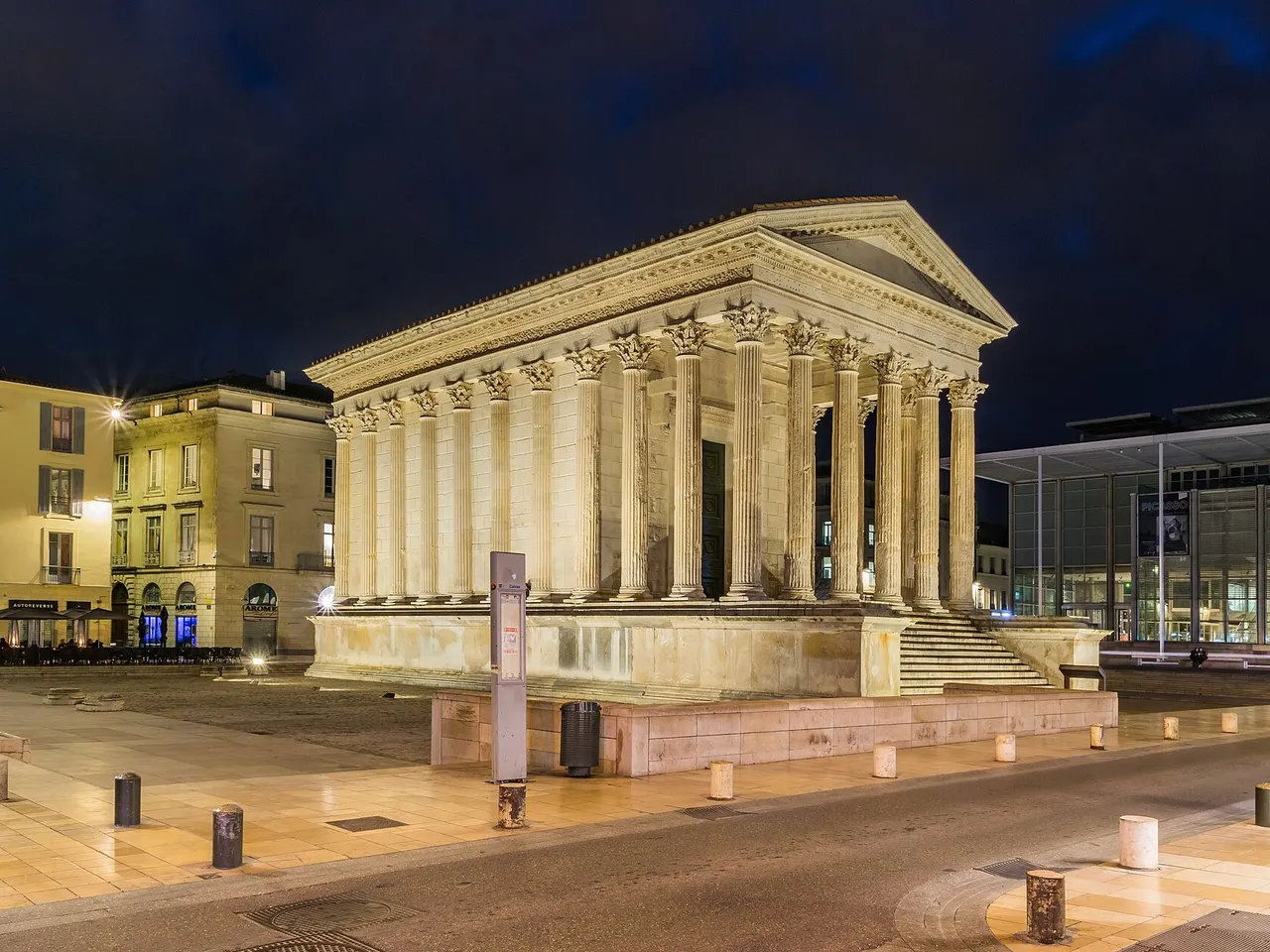 La Maison carrée de Nîmes illuminée la nuit, temple romain à colonnes corinthiennes, avec la façade vitrée du Carré d’Art sur la droite.