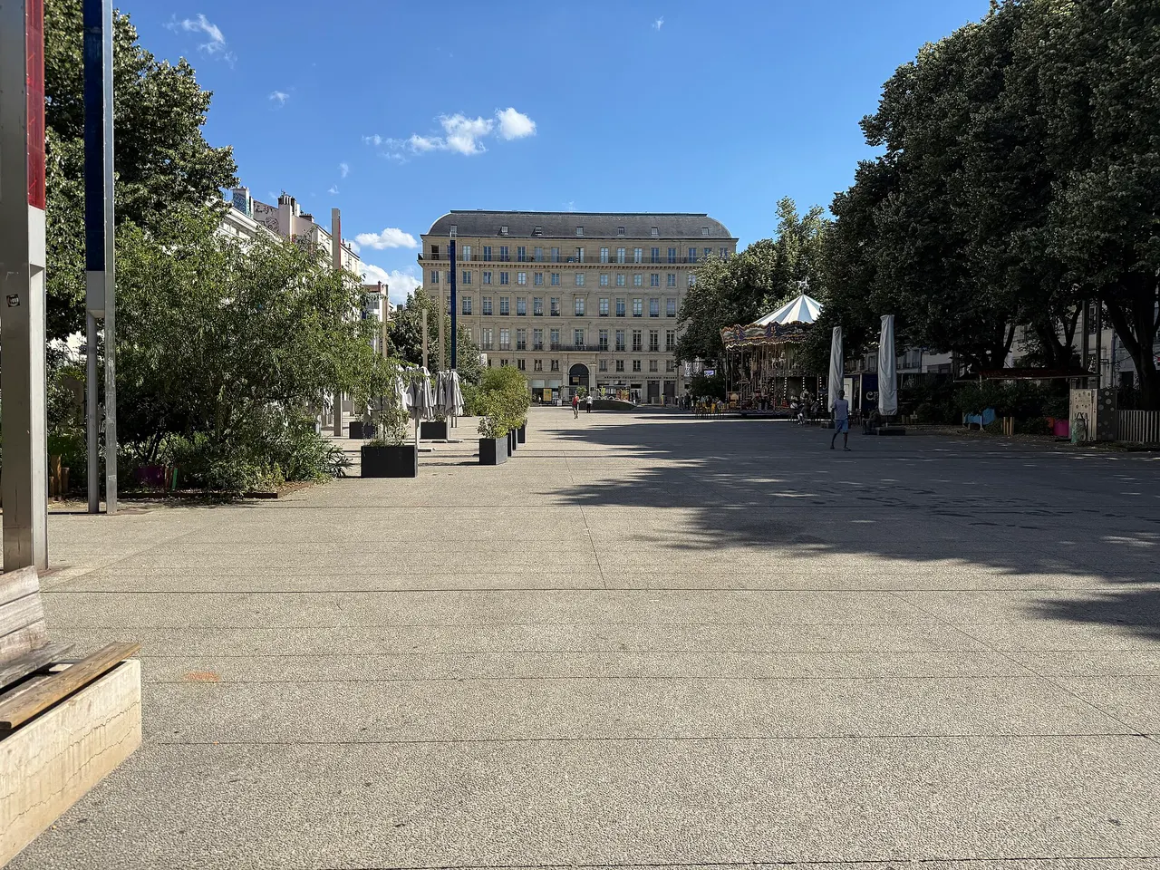La place de l’Hôtel-de-Ville de Saint-Étienne, piétonne et bordée d’arbres, avec un manège et un immeuble en pierre au fond