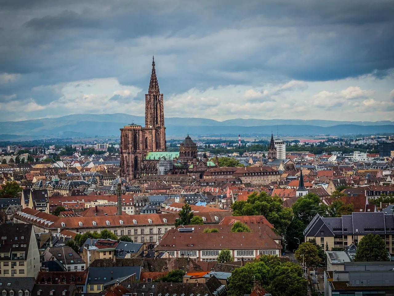La cathédrale Notre-Dame de Strasbourg et sa flèche de grès rose dominant les toits du centre, sous un ciel nuageux, montagnes à l’horizon.