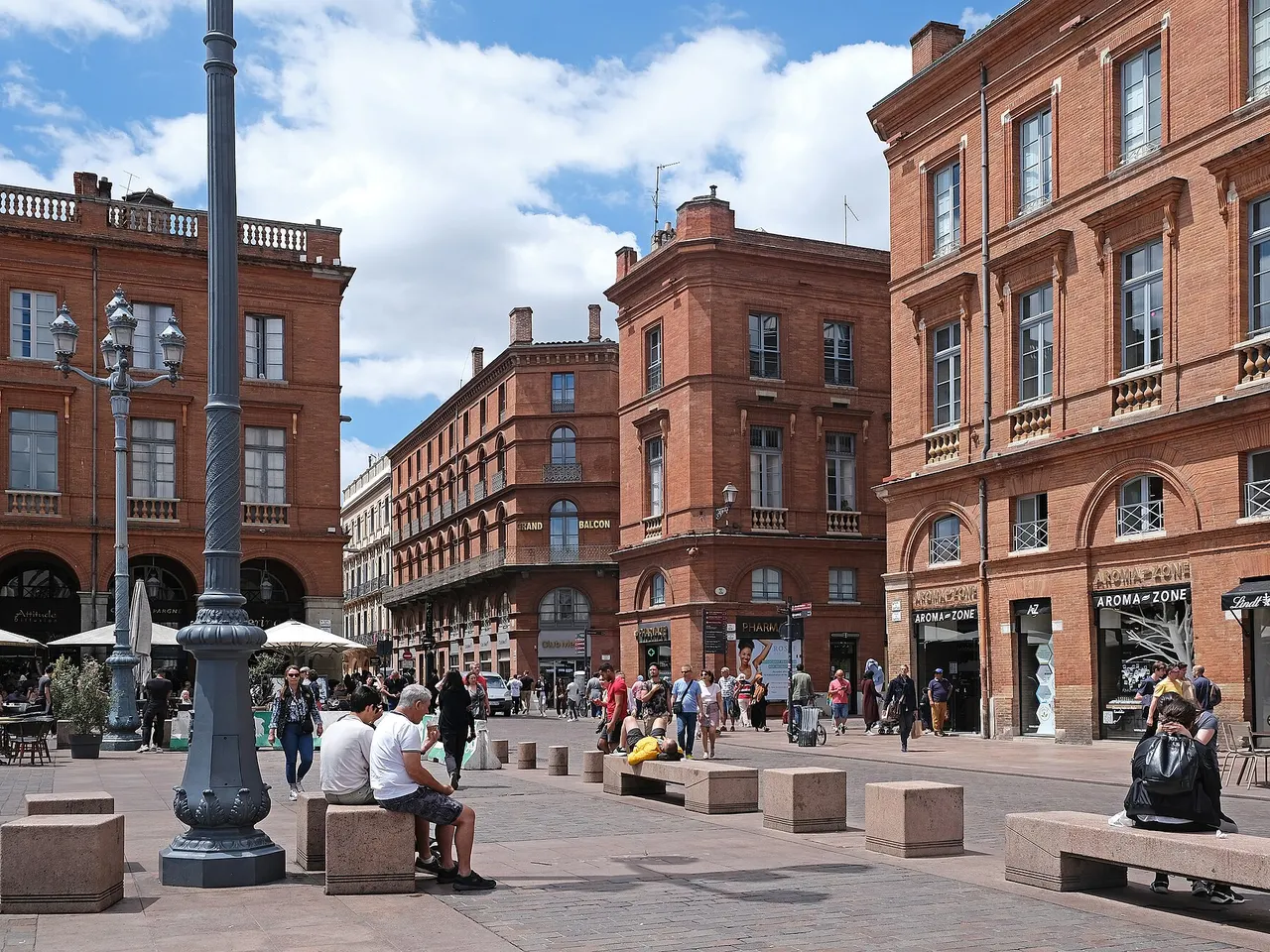 Façades de brique rose et passants sur une place piétonne du centre de Toulouse, aux abords de la place du Capitole.